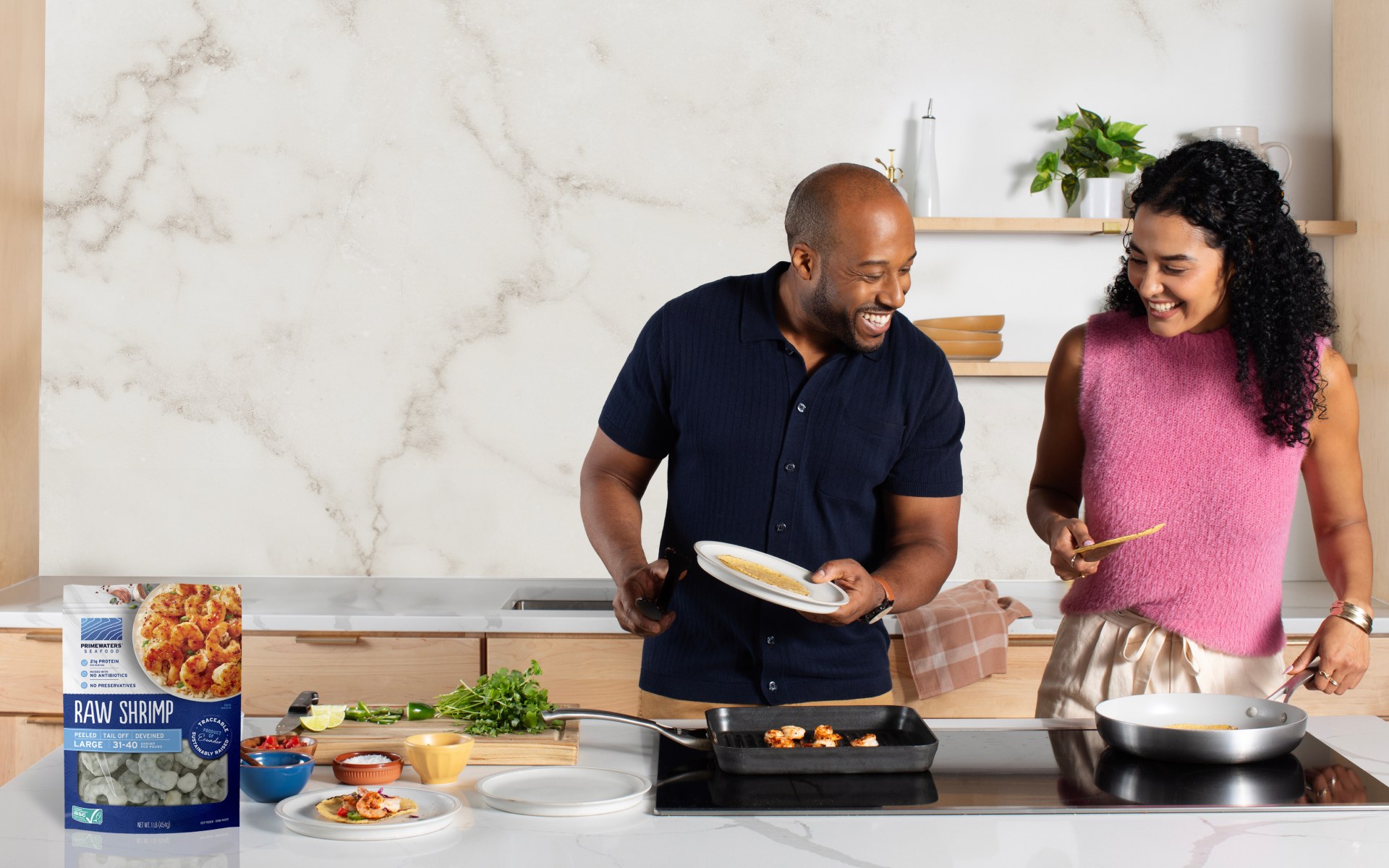 A couple cooking shrimp in a kitchen. A raw shrimp package is on the kitchen counter top